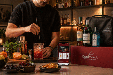 Here’s a clean, SEO-friendly **alt text** for your image: **Alt text:** A young adult crafting a cocktail at a cozy home bar using fresh ingredients and professional tools, with a Darumandu liquor delivery box, branded bottles, and a smartphone displaying the Darumandu app on a warmly lit kitchen counter at night. --- If you want a slightly more optimized version for SEO (blog use): **SEO alt text:** Home cocktail making scene with Darumandu online liquor delivery in Kathmandu, featuring a stylish bar setup, fresh ingredients, premium bottles, and a smartphone app interface in a warm, cozy kitchen environment.